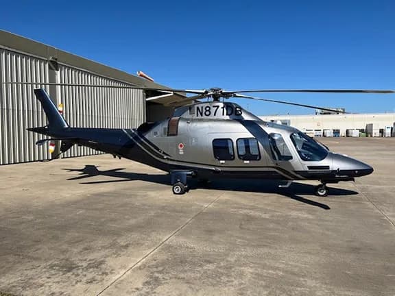 Side view of a silver and gray AgustaWestland AW109SP helicopter on the ground.