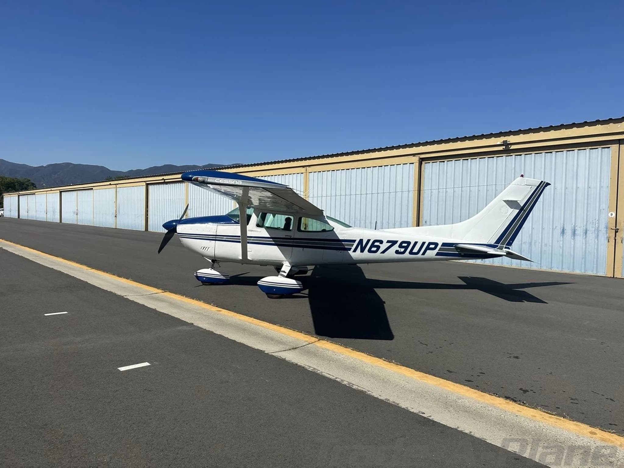 A white and blue Cessna 182 Skylane aircraft parked on an asphalt ramp with hangars and mountains in the background.