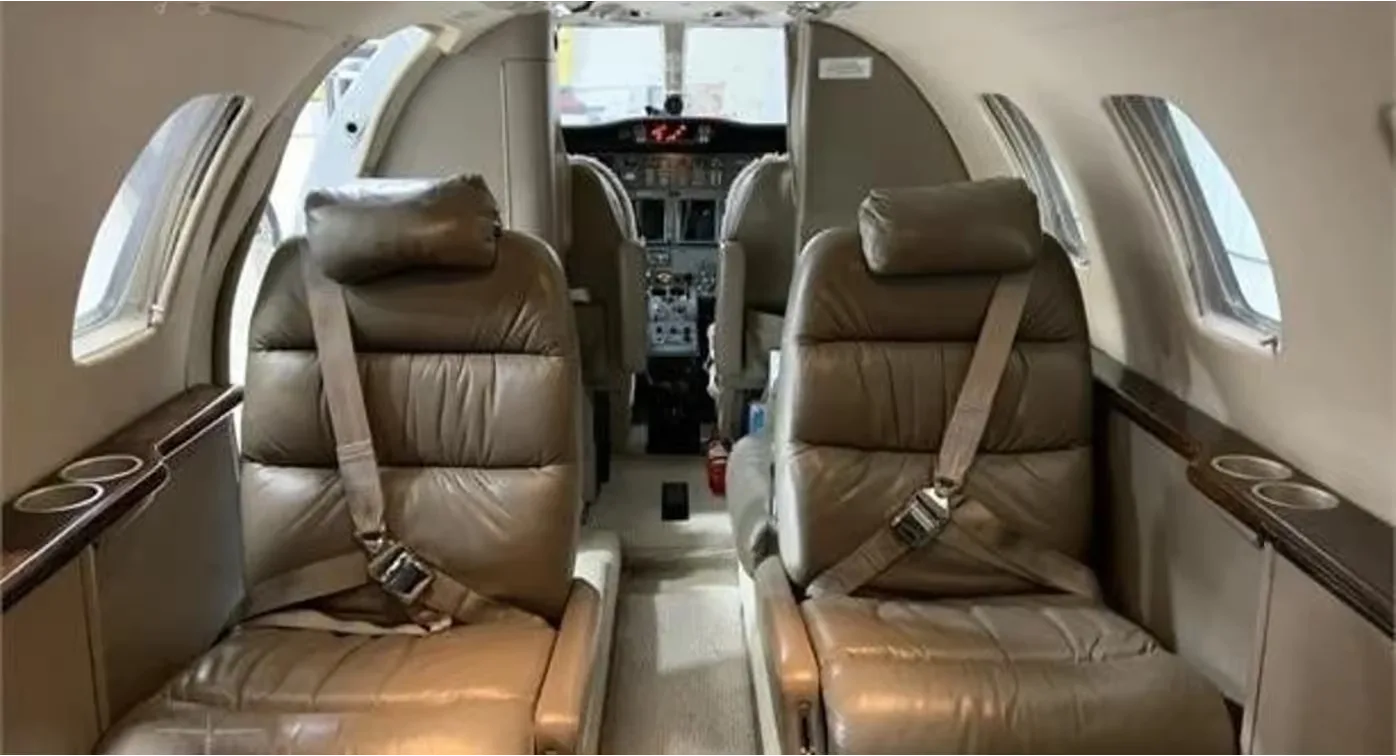 The aircraft cabin features two brown leather seats with seatbelts, looking towards the cockpit.