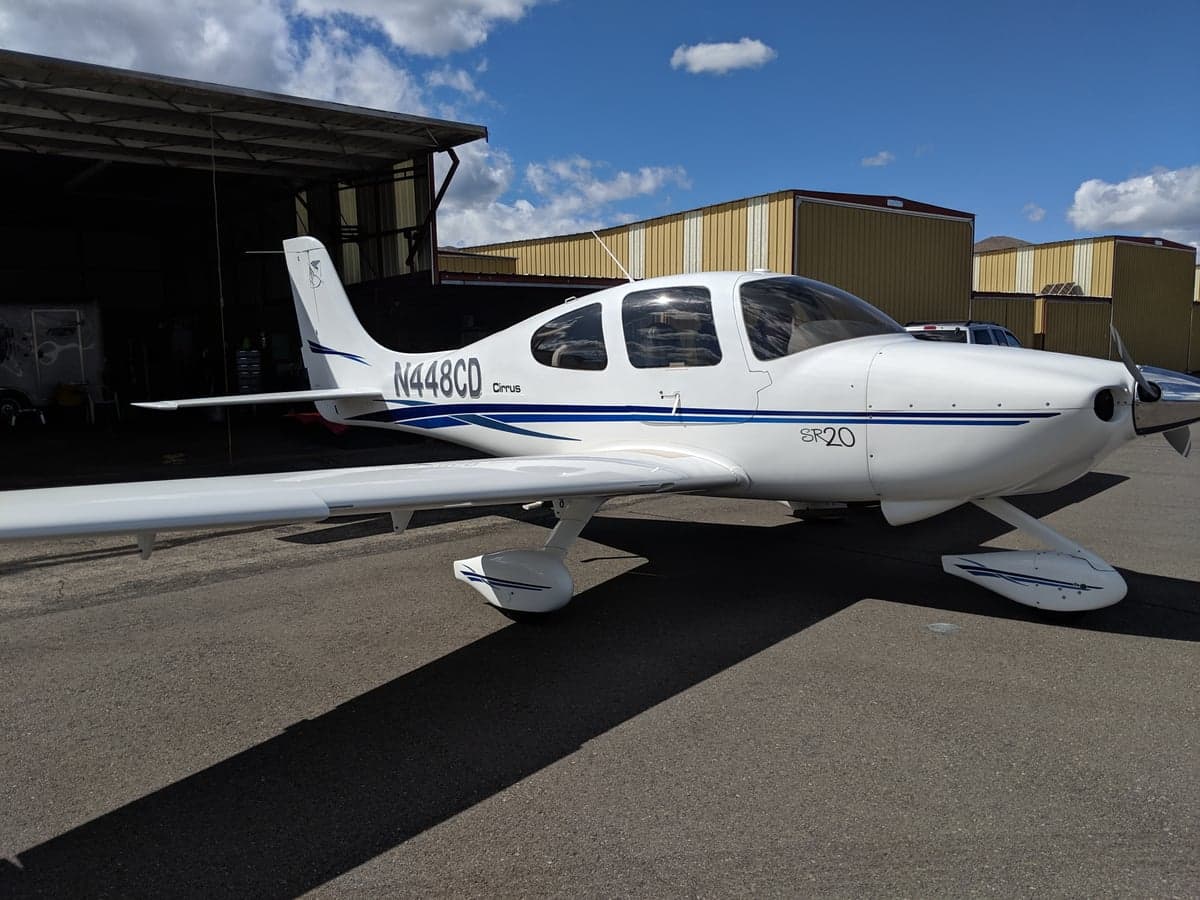 A white Cirrus SR20 aircraft with blue stripes (N448CD) parked on an asphalt ramp in front of a hangar under a partly cloudy sky.