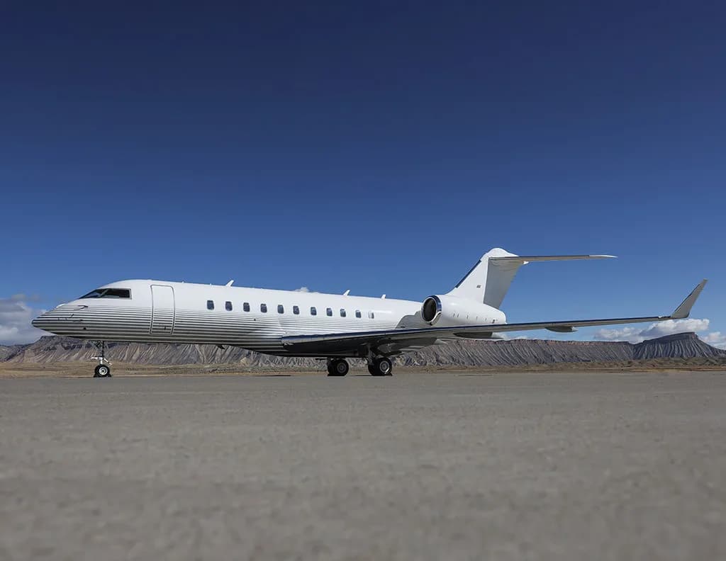 A white business jet with horizontal grey stripes on the fuselage is parked on an airfield with mountains in the background.