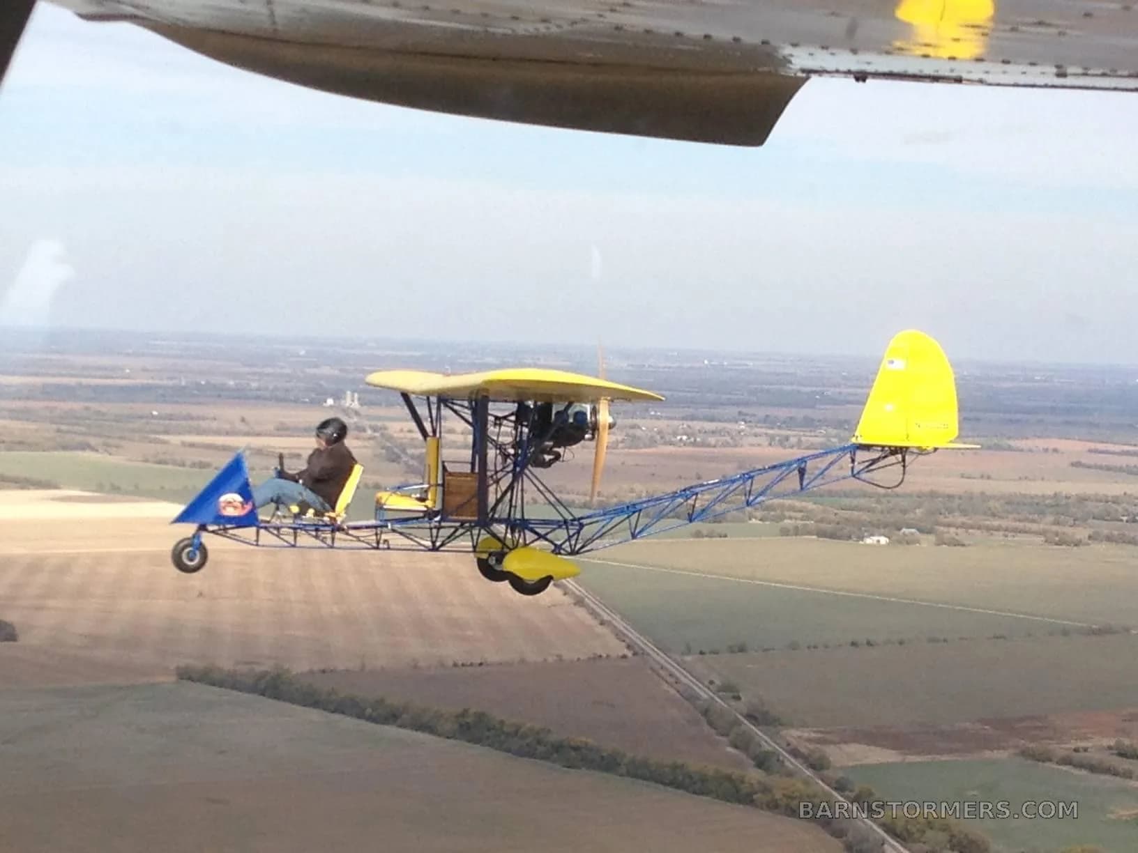 A yellow and blue ultralight aircraft with a single pilot flying over rural fields, viewed from another aircraft.