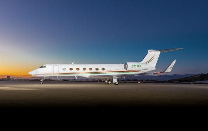 A white Gulfstream G550 business jet with green and red stripes is parked on a tarmac at dusk.