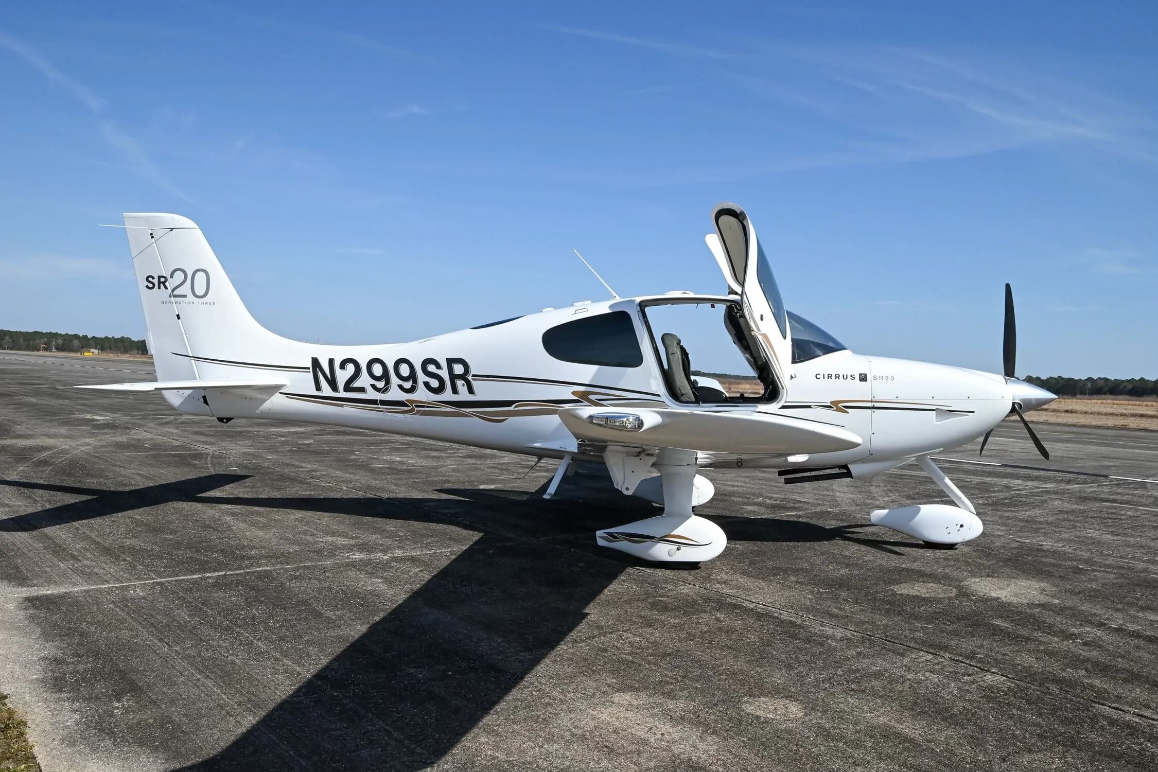A white Cirrus SR20 aircraft with black and gold accent stripes, registration N299SR, parked on an airfield with its right door open.
