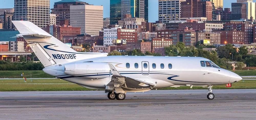 A white Hawker 800XP business jet with registration N900BF on a tarmac with a city skyline in the background.