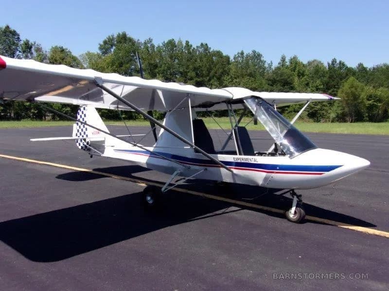 A white ultralight aircraft with blue and red stripes and a checkered tail, parked on an asphalt runway.
