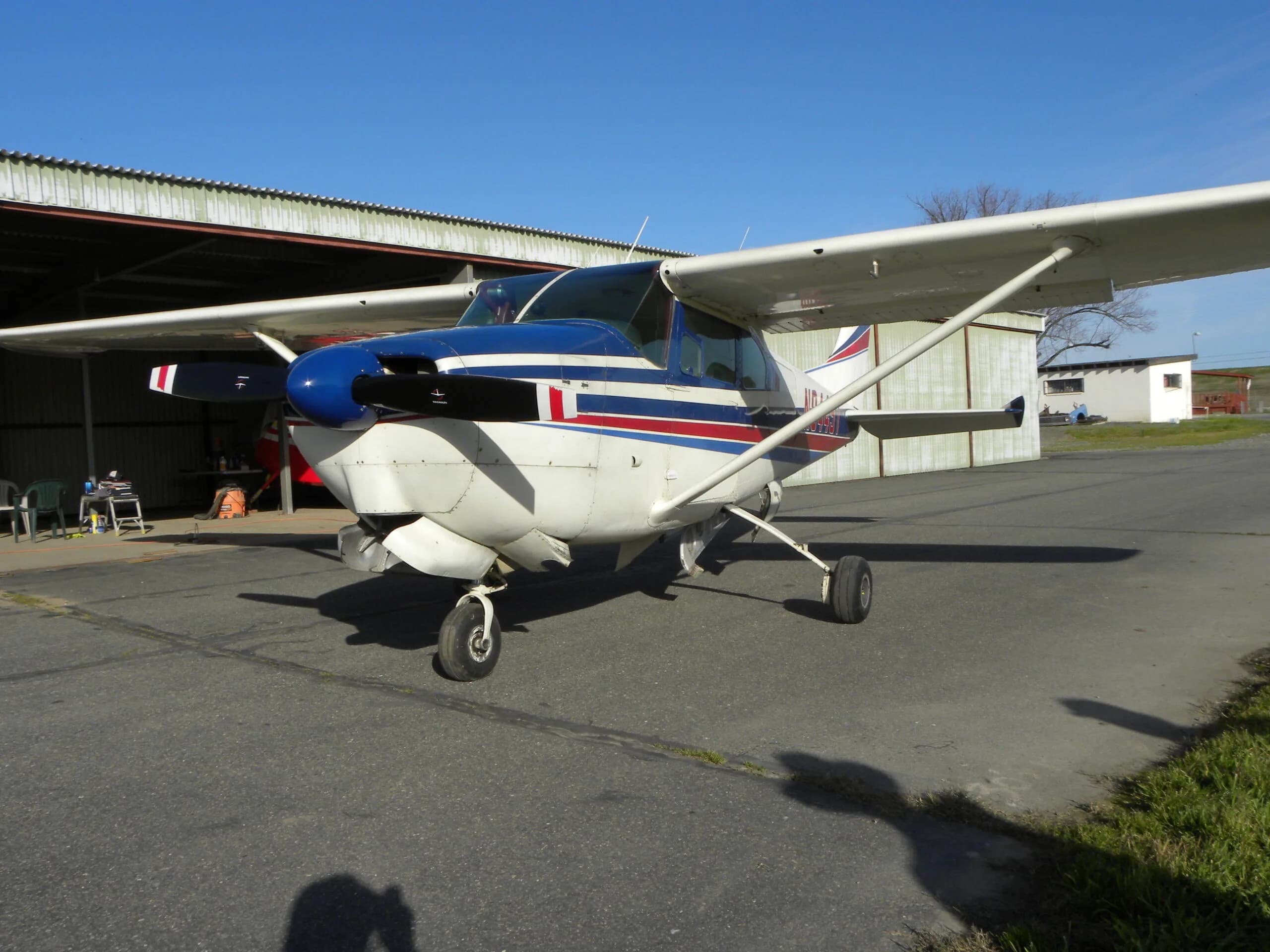 A front view of a white Cessna 182 with blue and red stripes, registration N9445T, parked in front of a hangar.
