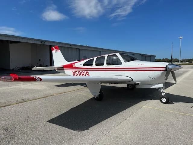 A white and red Beechcraft Bonanza N5861S parked on an asphalt ramp in front of a hangar under a blue sky.
