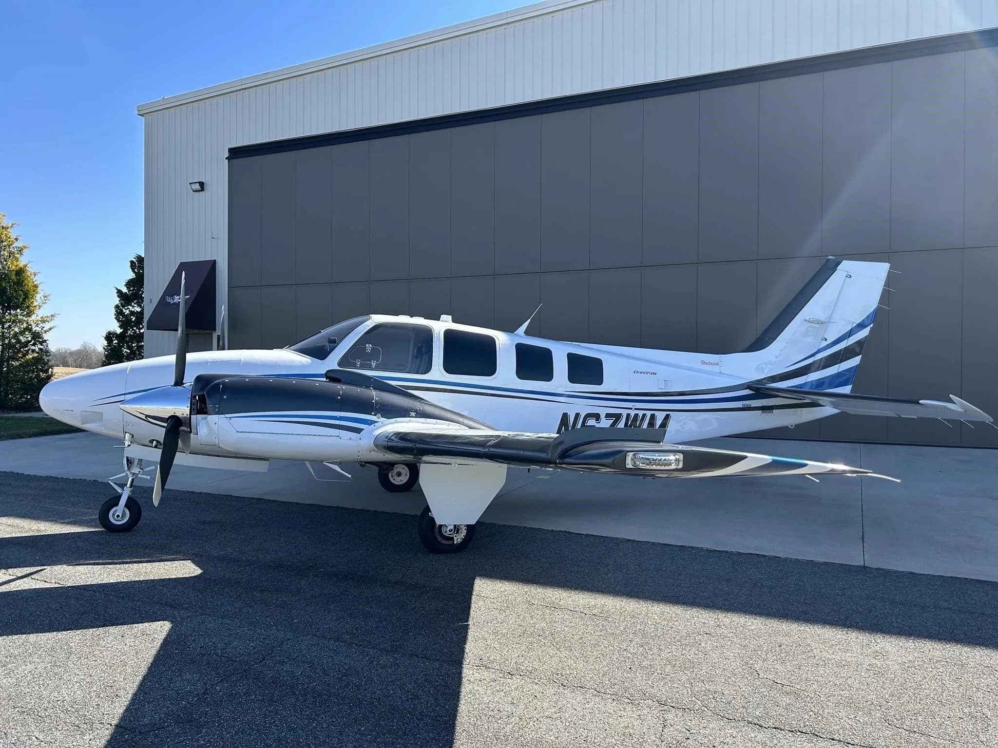 A white and blue Beechcraft Baron N67WM parked on an tarmac outside a hangar on a sunny day.