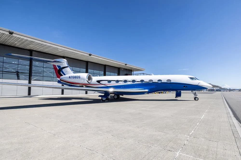 An aerial view of a Gulfstream G650 business jet with a white and blue paint scheme, parked on an airport runway.