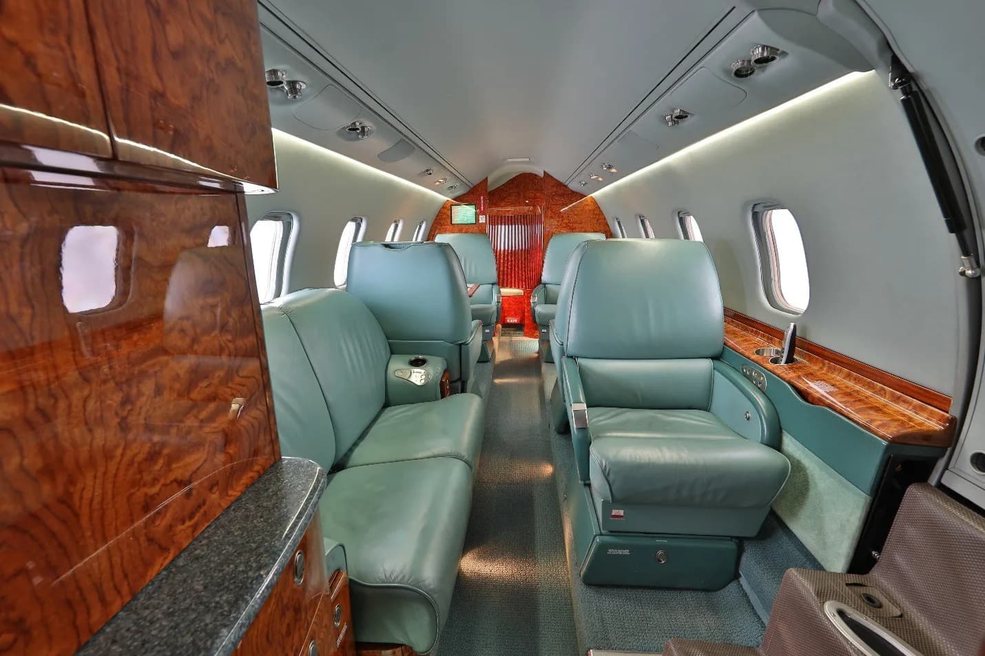 A view down the aisle of a private jet's cabin, showing teal leather seats, polished wood cabinetry, and windows.