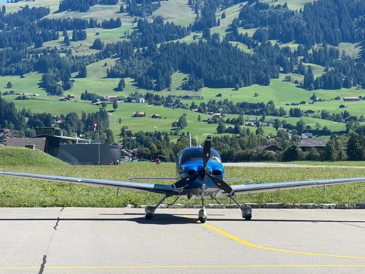 A blue and silver Cirrus SR22 aircraft parked on an airfield with lush green mountains and houses in the background.