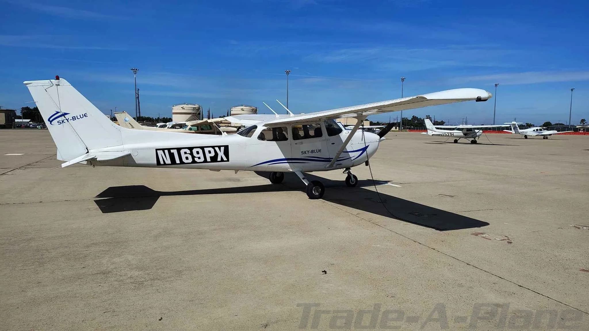 A side view of the white Cessna 172, N169PX, with blue accents, parked on an airport tarmac.