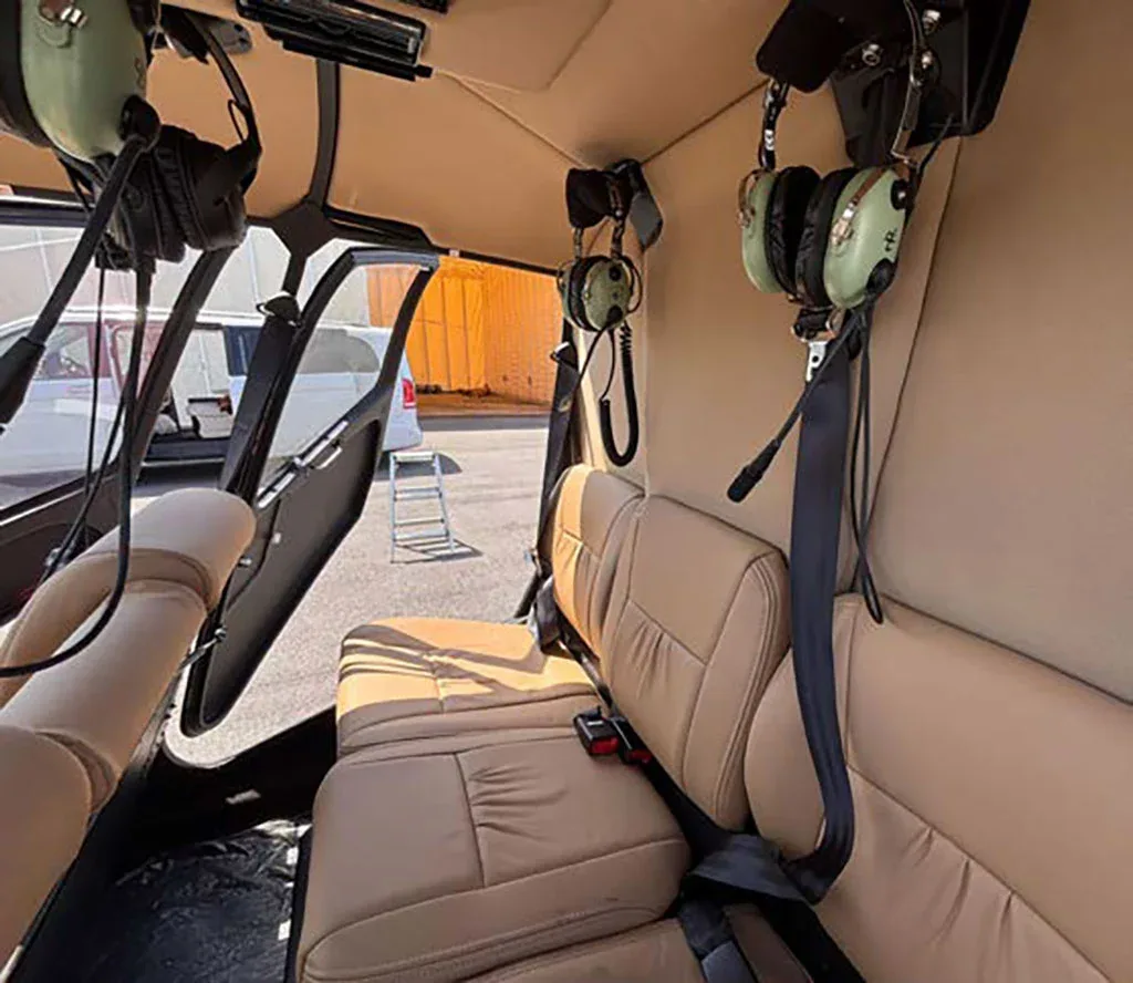 The tan leather interior of a helicopter cabin is visible, with headsets hanging and an open door revealing the exterior.