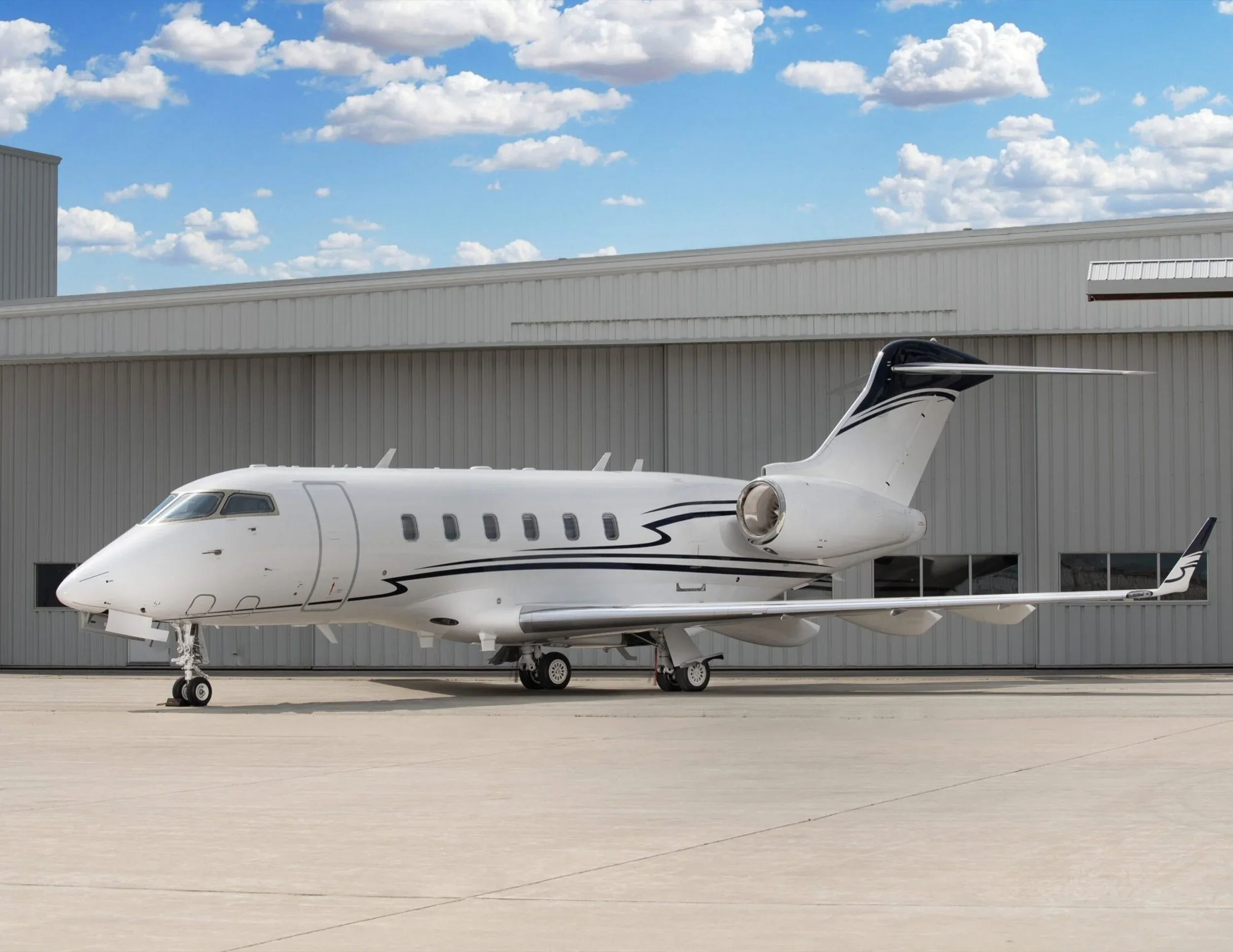 A white Bombardier Challenger business jet with a dark blue stripe on the fuselage and tail, parked on a tarmac in front of a hangar.
