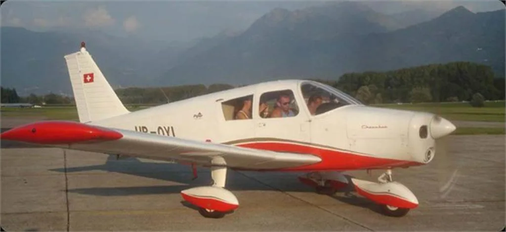 A white and red Piper Cherokee aircraft (HB-OYI) with people inside, parked on an airfield with mountains in the background.
