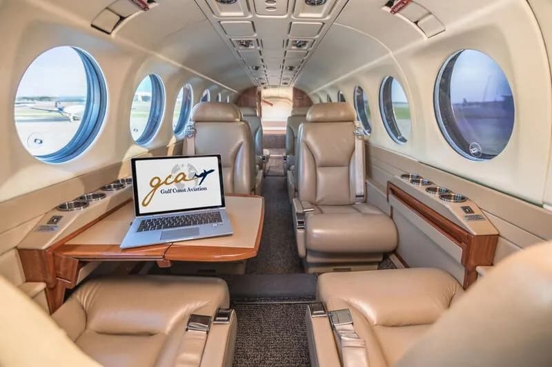 The aircraft cabin features tan leather seats, a small table with a laptop displaying a logo, and round windows.
