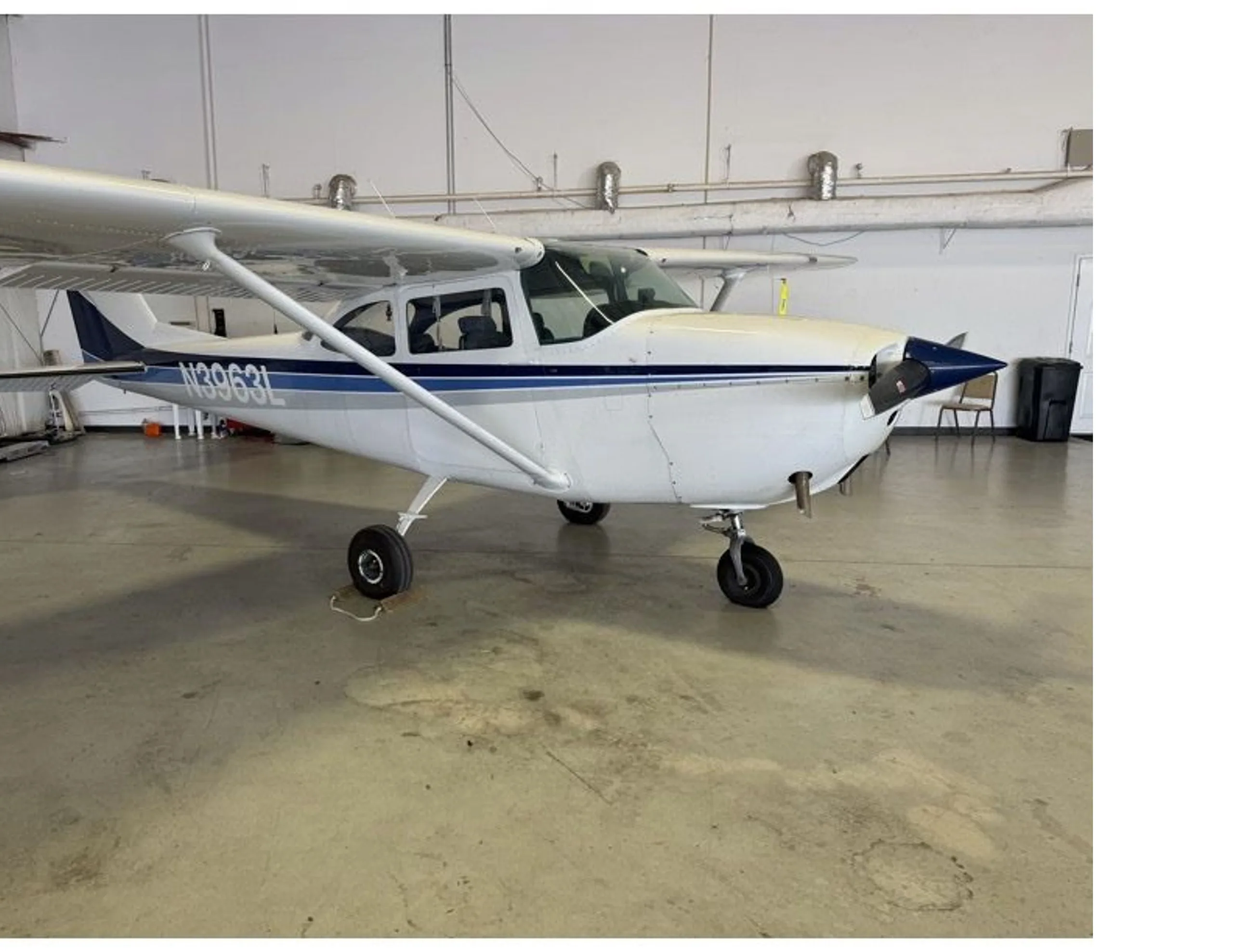 A front-side view of a white Cessna 172 with blue and grey stripes, registration N3963L, parked in a hangar.