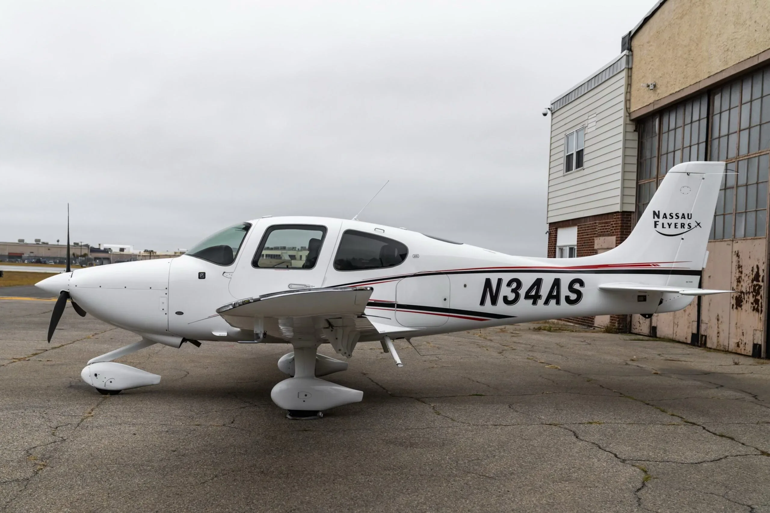 A white Cirrus SR22 aircraft with red and black stripes, registration N34AS, parked on an airfield next to a building.