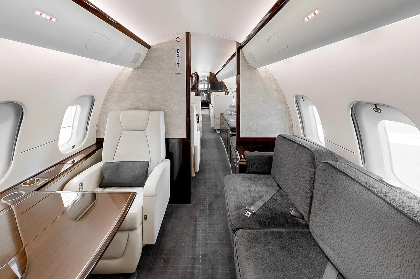 The luxurious interior cabin of a business jet, featuring a grey sofa, a cream armchair with a table, and dark wood accents.