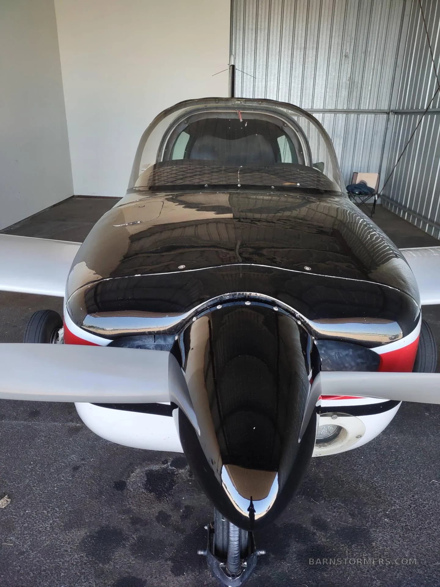 A head-on view of the propeller and nose of a white, red, and black single-engine aircraft inside a hangar.