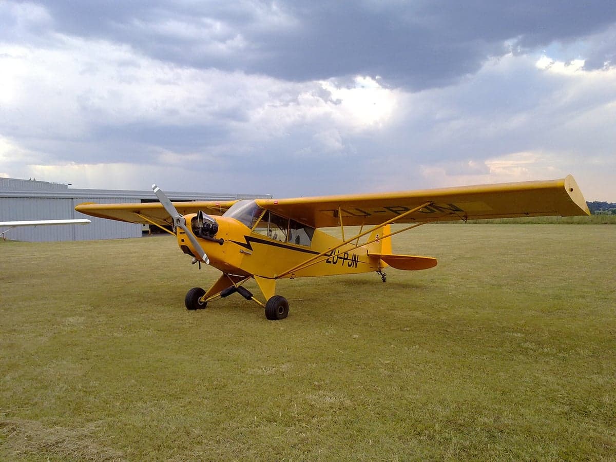 A yellow Piper Cub sits on a grassy airfield under a cloudy sky.