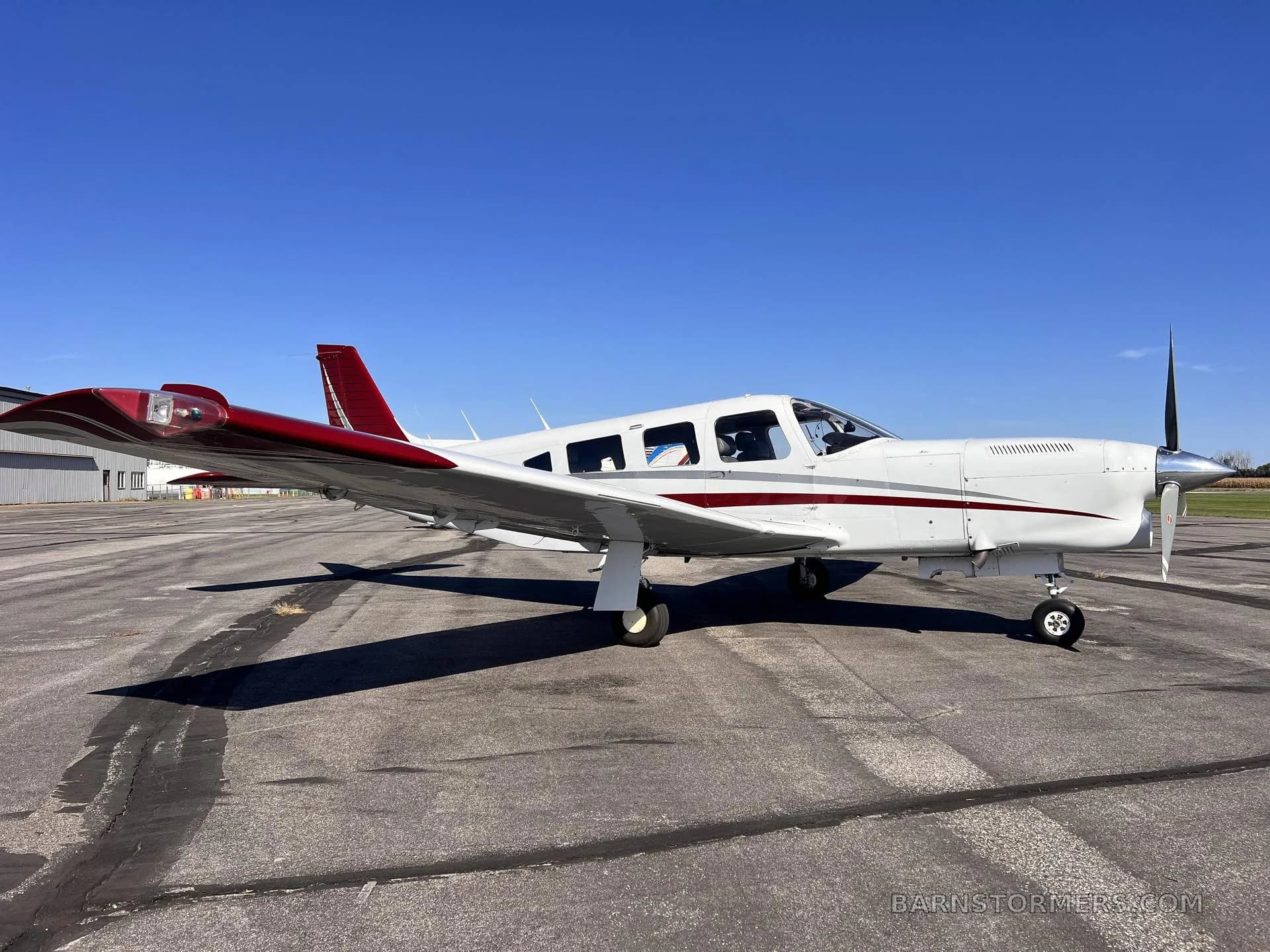 A white single-engine propeller aircraft with red and white tail and stripes, parked on an asphalt ramp under a clear blue sky, viewed from the front-right.