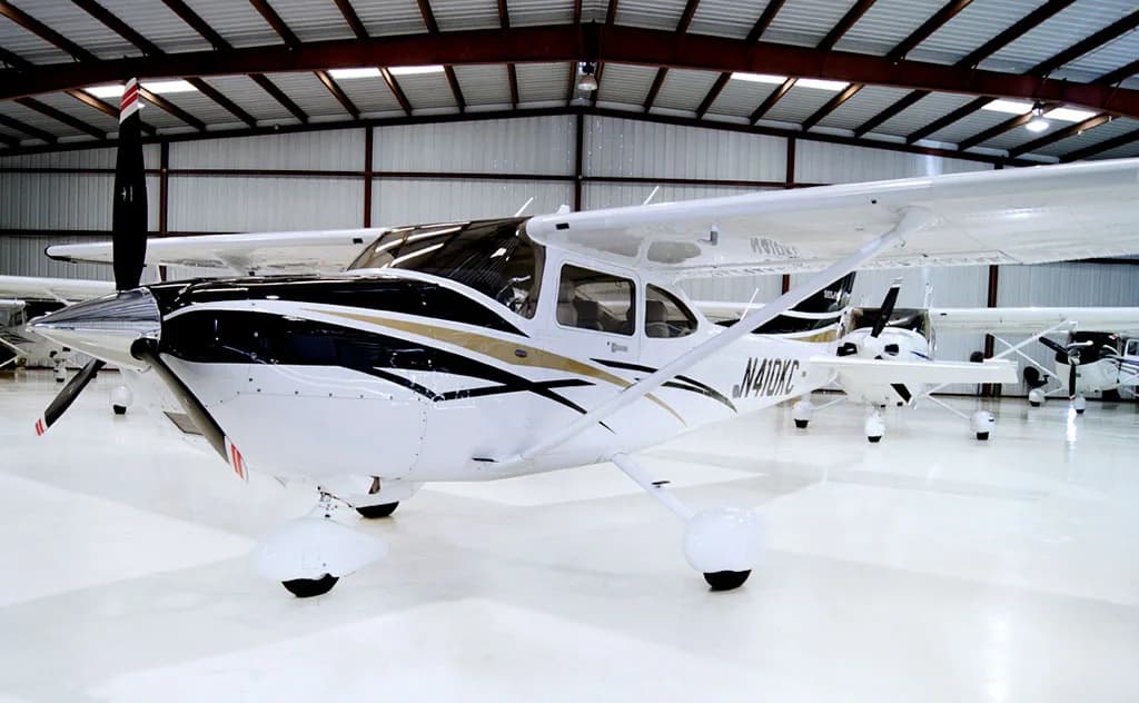 A white Cessna Skylane aircraft with black and gold stripes is parked in a hangar, viewed from the front-left.
