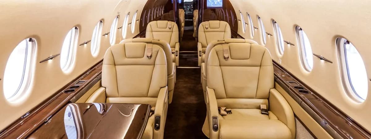A view down the aisle of a luxurious aircraft cabin with tan leather seats and dark wood accents.