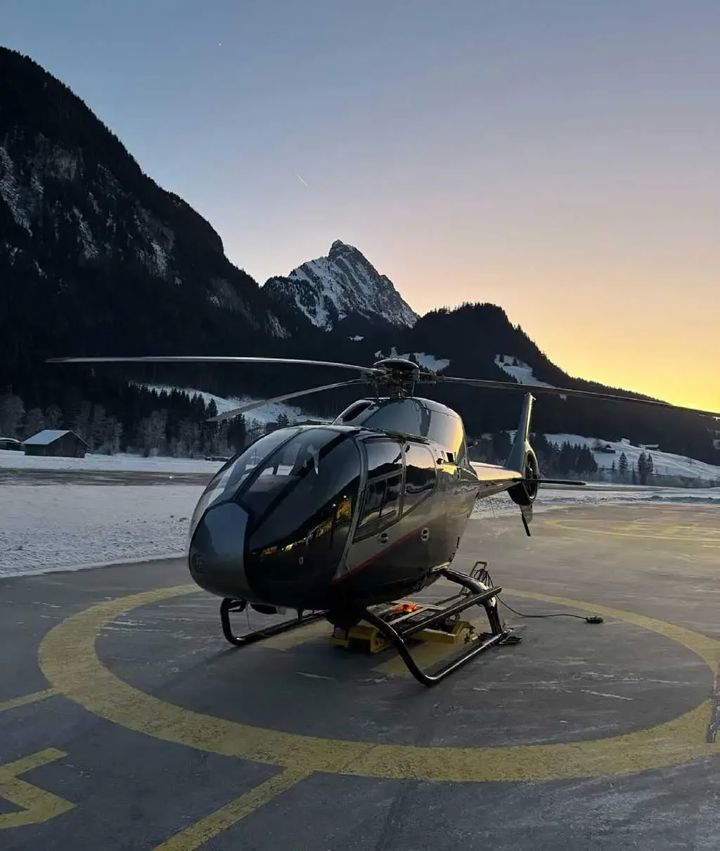 A dark grey helicopter is parked on a helipad at sunset, surrounded by snow-covered mountains.