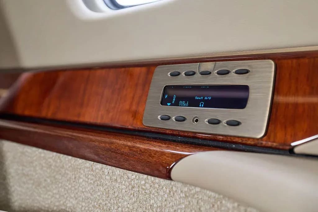 A close-up of a brushed metal seat audio/video control panel embedded in the polished wood side panel of the cabin.