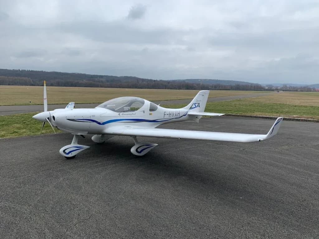 A white Dyn'Aéro MCR01 Hummingbird light aircraft with blue stripes is parked on an airfield tarmac under a cloudy sky.