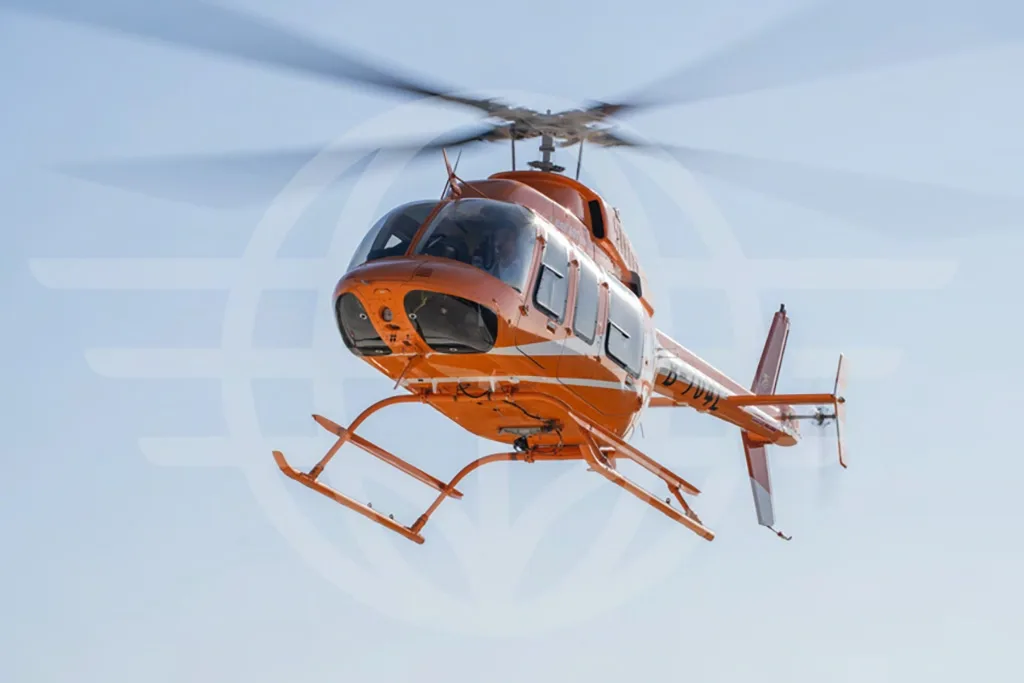 An orange Bell 407 helicopter with white stripes in flight, viewed from the front-right.