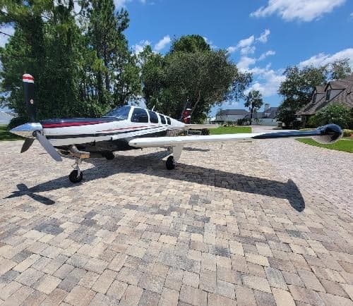 A Beechcraft Bonanza sits on a brick driveway in front of a house and trees.