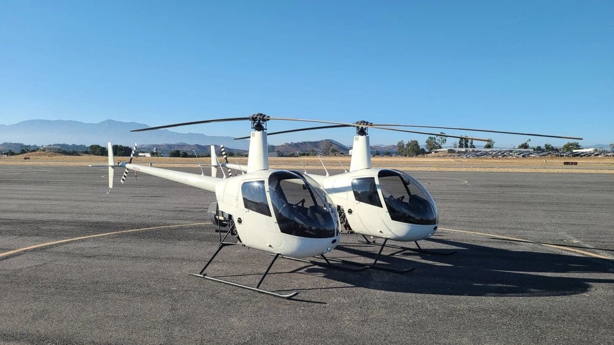 Two white Robinson R22 helicopters are parked side-by-side on an airport tarmac under a clear blue sky.