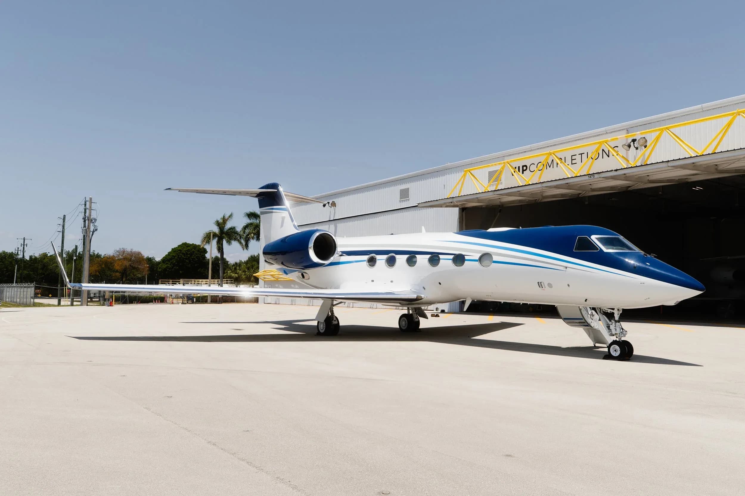 A Gulfstream G550 business jet with a blue and white paint scheme parked on an airfield next to a hangar.