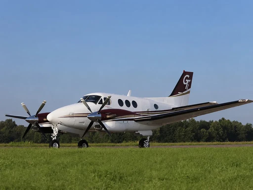 A white and maroon twin-turboprop aircraft with a 'GT' logo on its tail sits on a grassy airfield under a clear blue sky.