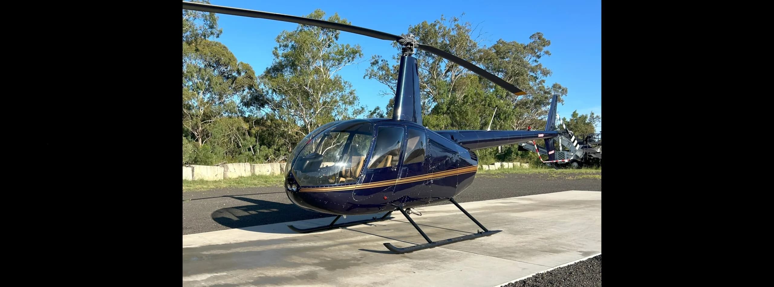 A dark blue Robinson R44 helicopter with gold stripes is parked on a concrete pad under a clear sky.