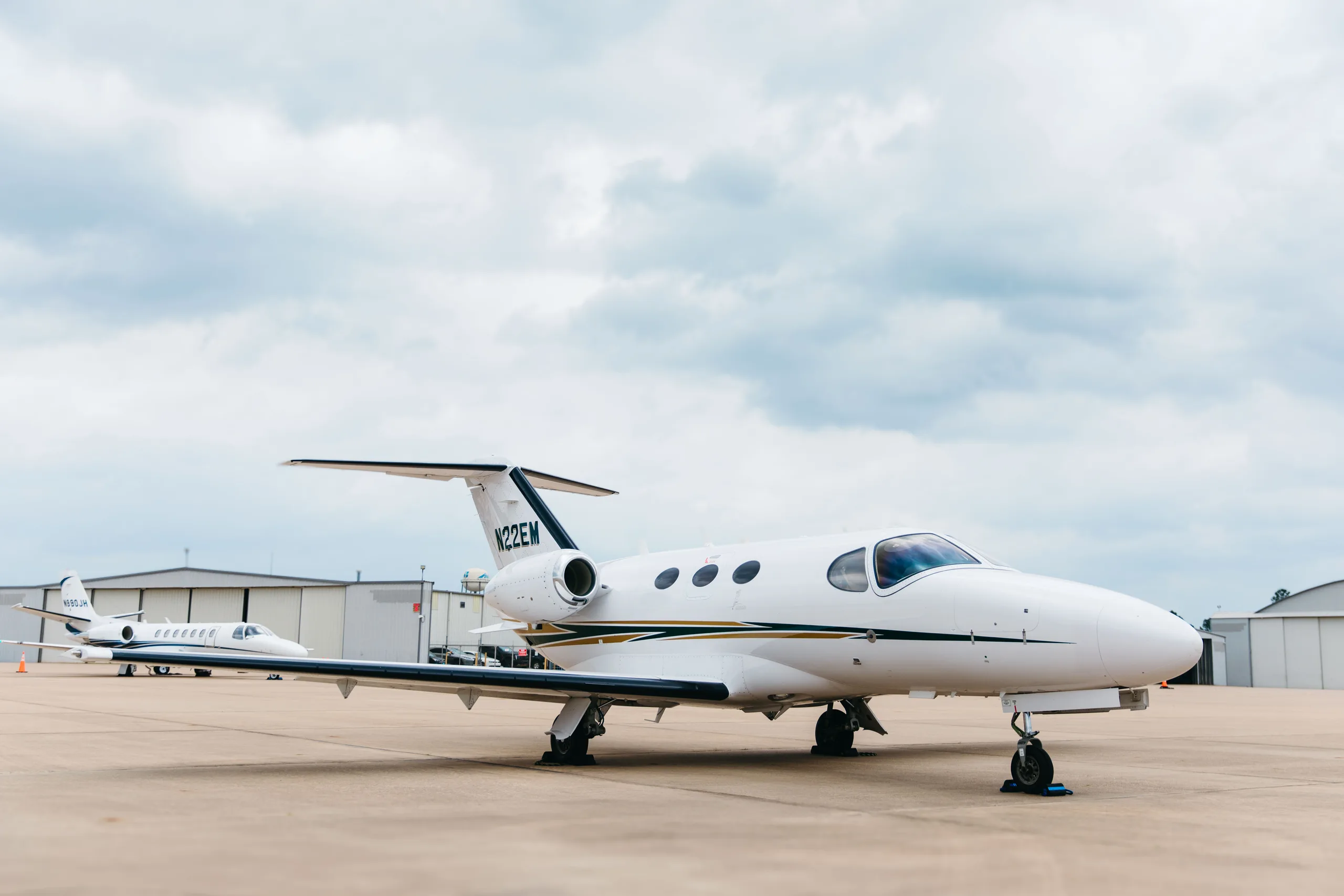 A three-quarter front view of a white Cessna Citation M2 business jet, tail number N22EM, with gold and dark green stripes, parked on a tarmac with another jet.