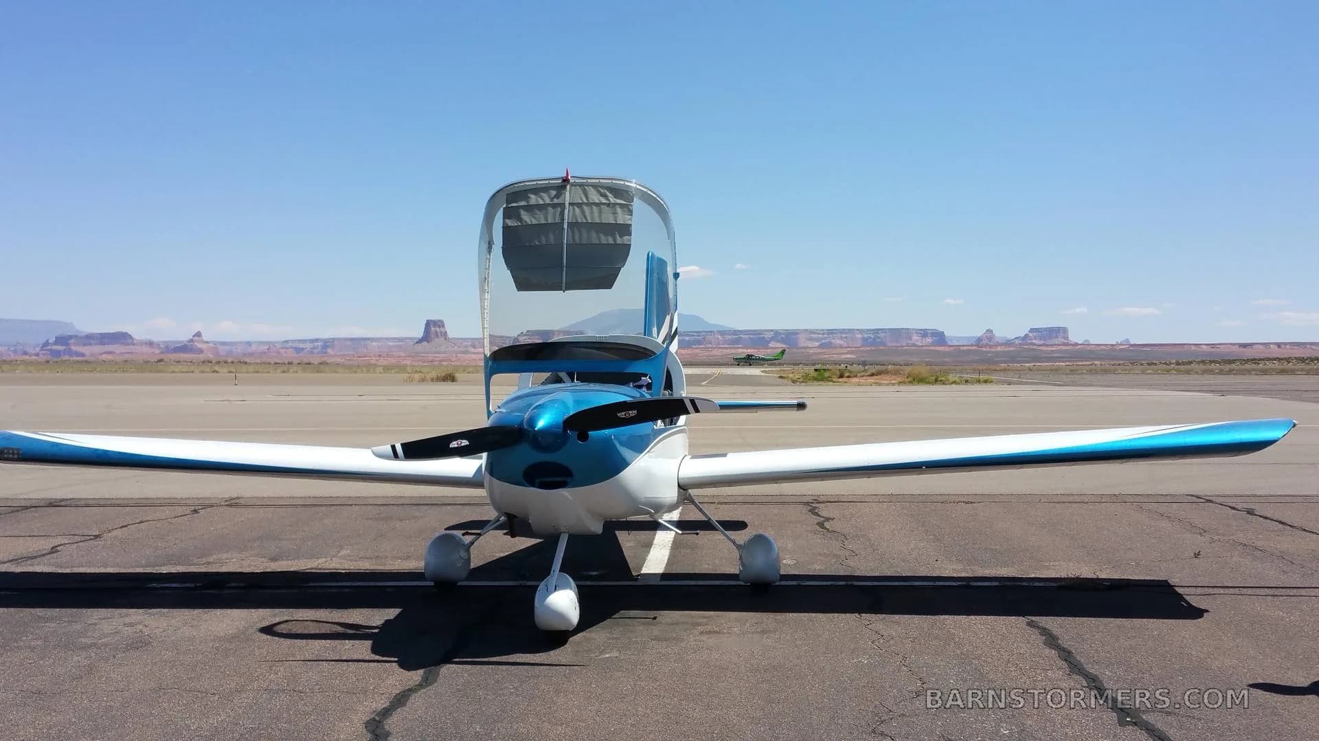 A blue and white single-engine aircraft with its canopy open is parked on an airfield under a clear blue sky with distant mesas.