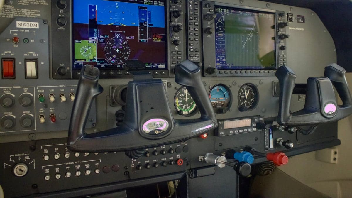 The cockpit of an aircraft, showing two yokes and a glass panel display with flight information.