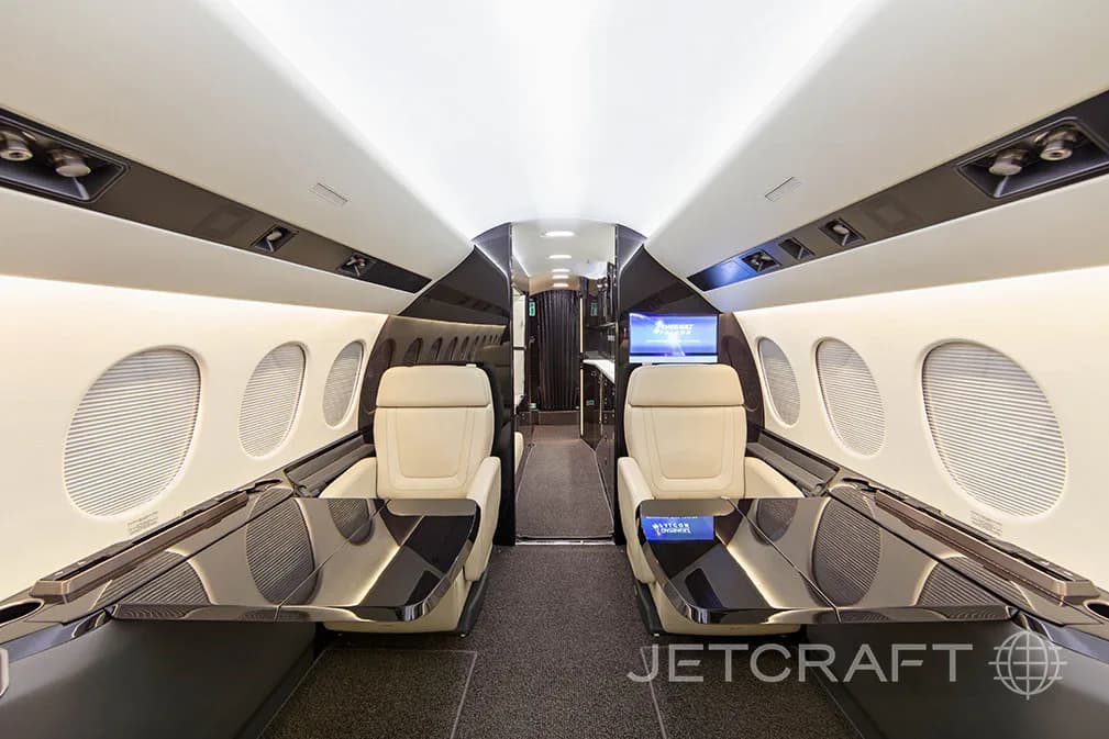 The spacious interior of a private jet cabin, featuring two cream-colored seats facing each other with dark glossy tables.