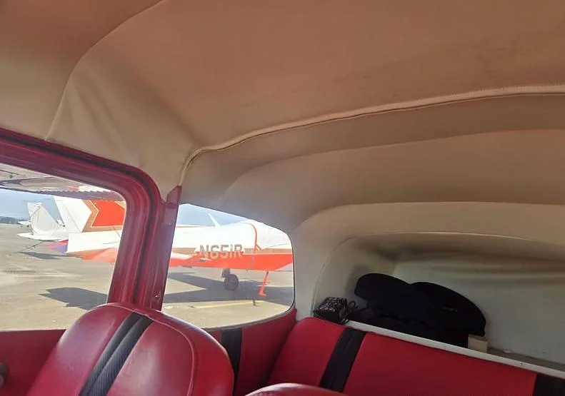 The red and tan interior of an aircraft cabin, looking out the window at another red and white aircraft on the tarmac.