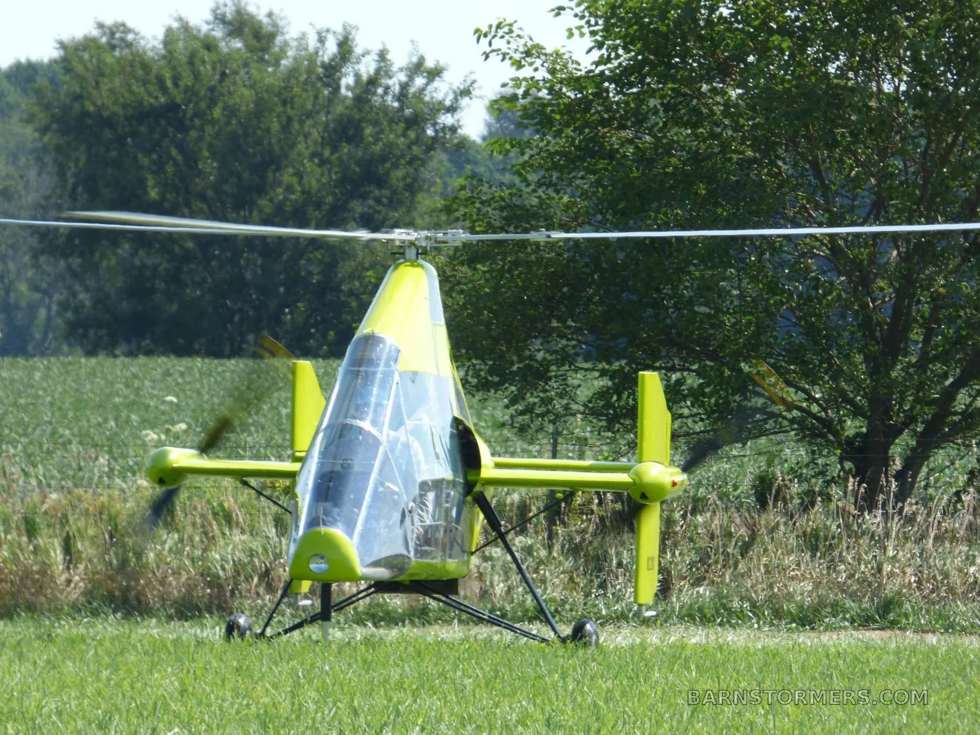A bright green gyrocopter with a clear canopy, seen from the front, parked on a grassy field with trees in the background.