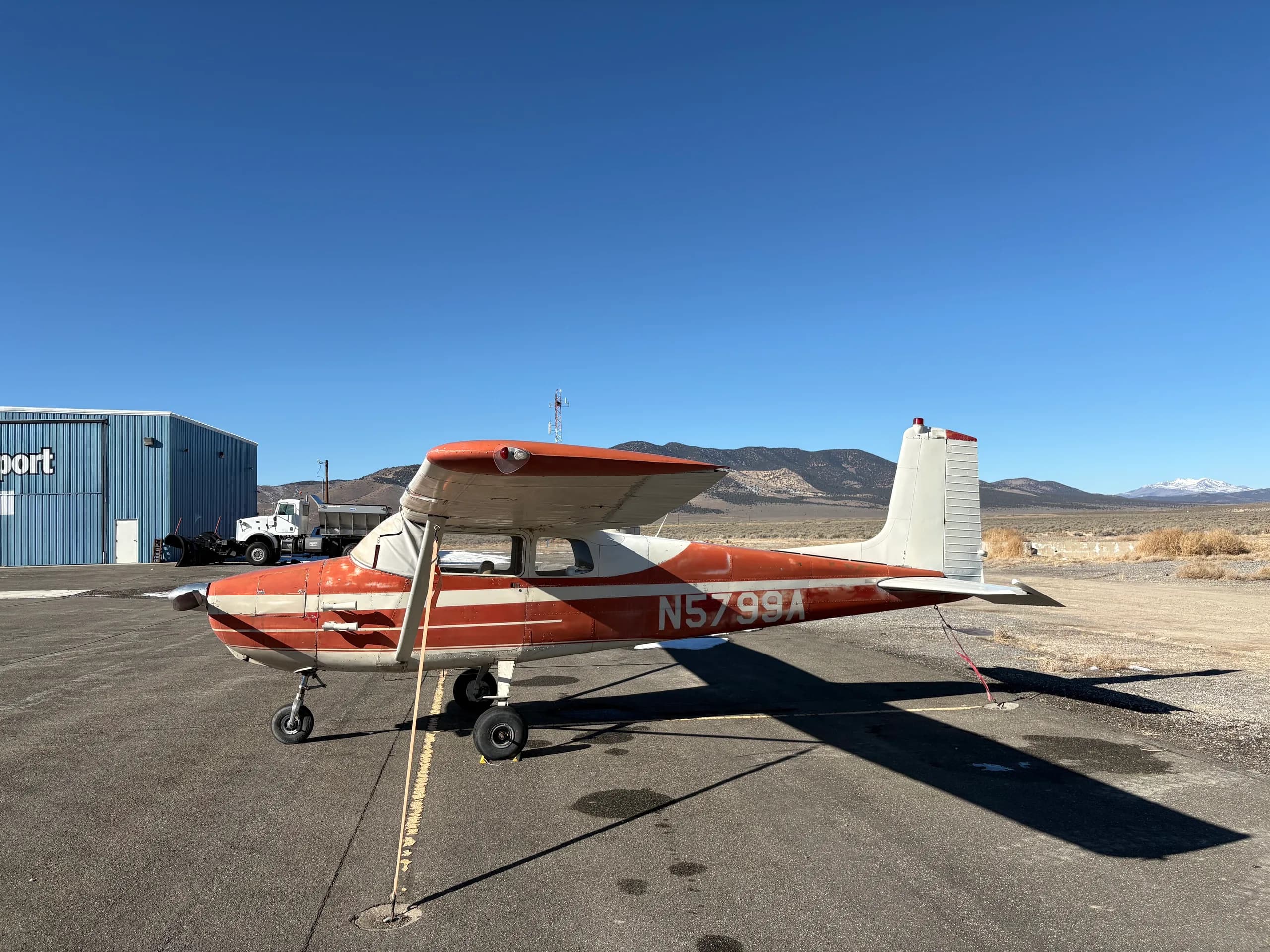 A red and white single-engine high-wing aircraft, N5799A, parked on an airfield with a blue hangar and mountains in the background.