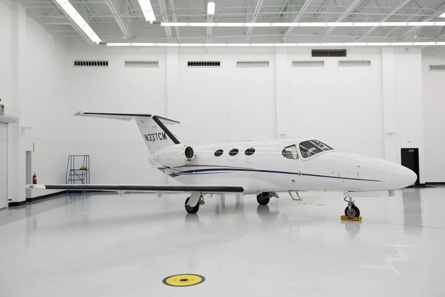 A white Cessna Citation M2 business jet with blue and gold stripes, tail number N337CM, parked in a brightly lit hangar.