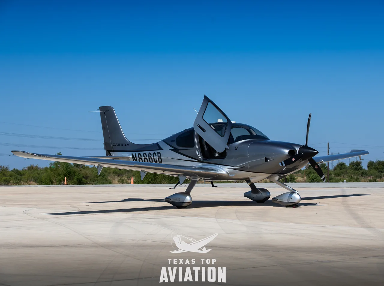 A three-quarter front view of a silver and grey Cirrus SR22 aircraft with registration N886CB and open doors.
