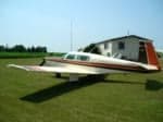 A white single-engine propeller aircraft with red stripes and an orange tail is parked on a grassy field with a building in the background.