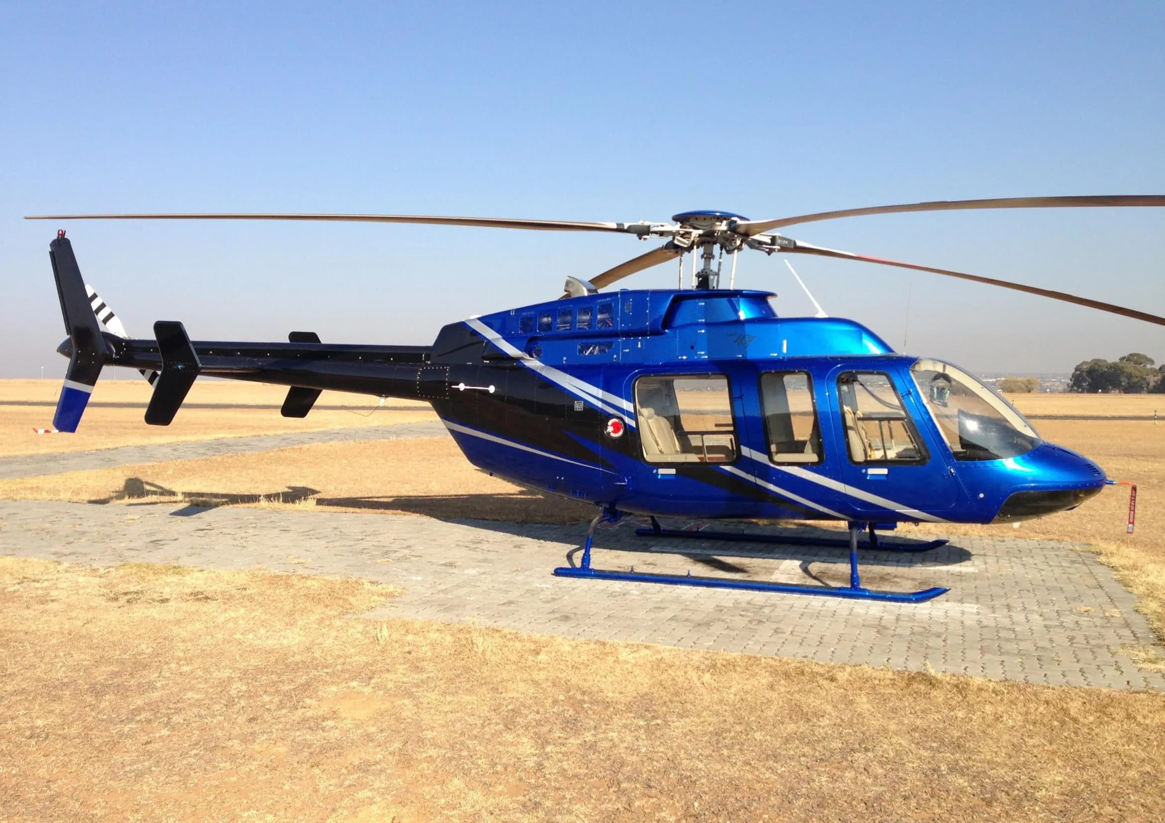 A blue Bell 407 helicopter with black and white stripes is parked on a paved area in a dry, grassy field under a clear sky.