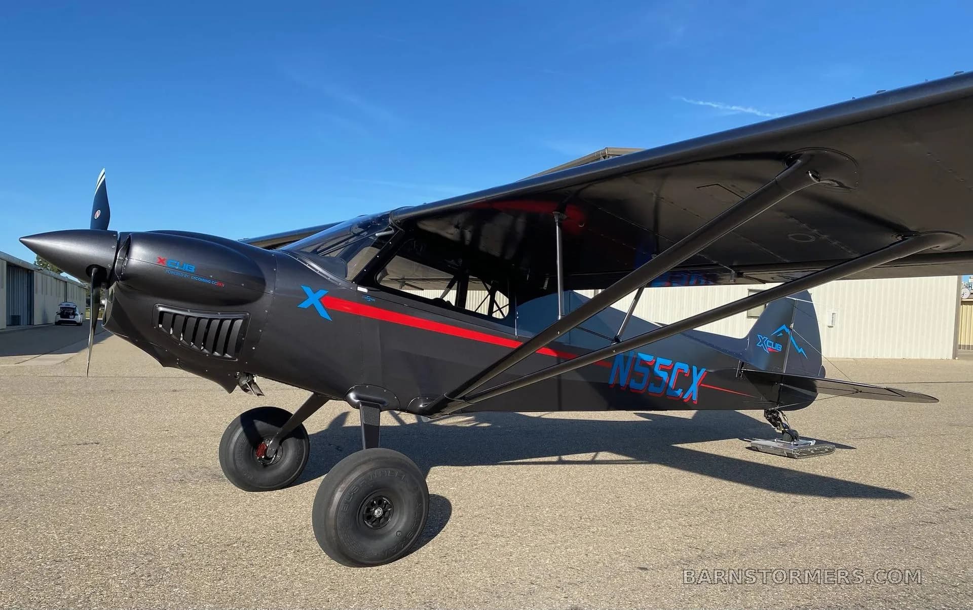 A dark gray X-Cub aircraft with red and blue accents, registration N55CX, parked on an asphalt ramp with hangars in the background.